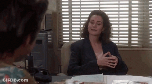 A Woman In A Suit Is Sitting At A Desk In Front Of A Window . GIF