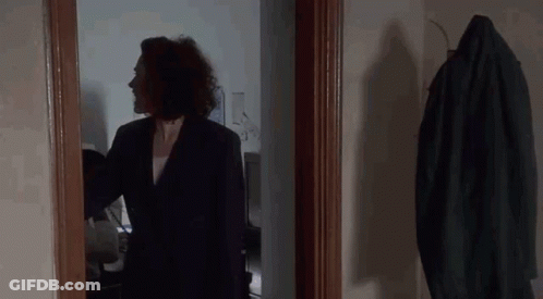 A Woman In A Suit Is Standing In A Hallway Next To A Coat Hanging On A Rack . GIF