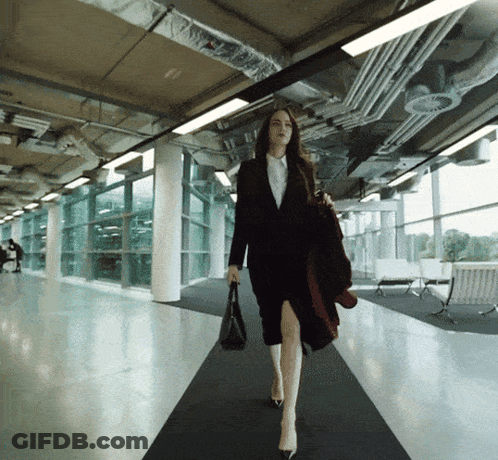 A Woman In A Suit Is Walking Down A Hallway Holding A Purse GIF