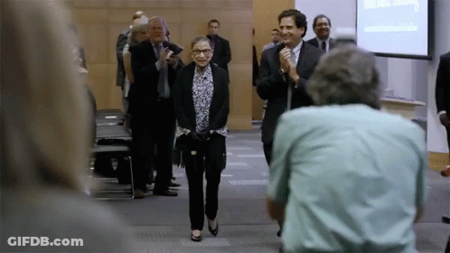 A Woman In A Suit Is Walking Down A Hallway With A Man Holding A Microphone Behind Her GIF