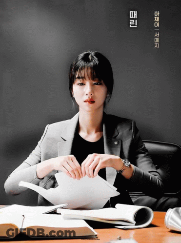 A Woman In A Suit Sits At A Desk Holding A Piece Of Paper In Front Of A Poster With Chinese Writing On It GIF