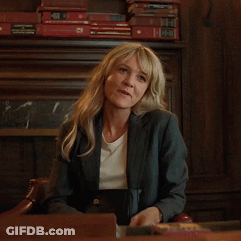 A Woman In A Suit Sits At A Desk With A Stack Of Books Behind Her That Says ' A ' On It GIF