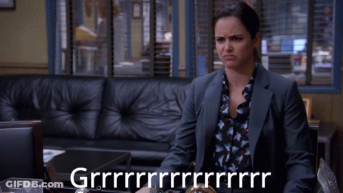 A Woman In A Suit Sits In Front Of A Couch And A Sign That Says Grrrr GIF