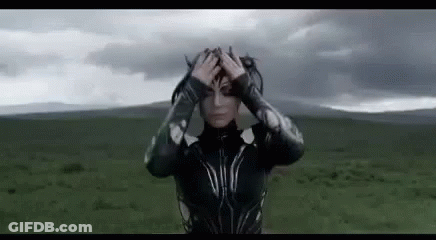 A Woman In A Superhero Costume Is Standing In A Field Holding Her Head . GIF