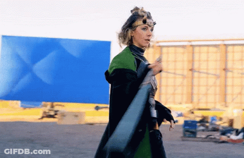 A Woman In A Superhero Costume Is Standing In Front Of A Blue Wall Holding A Sword . GIF