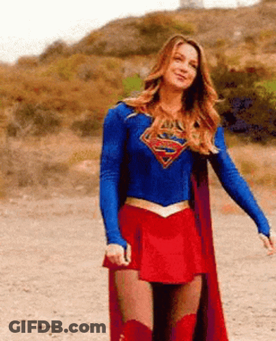 A Woman In A Superman Costume Is Standing In A Dirt Field . GIF