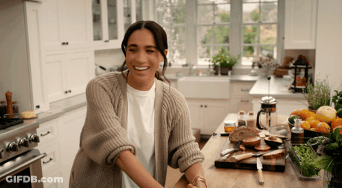 A Woman In A Sweater Is Smiling While Standing In A Kitchen GIF