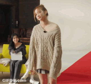 A Woman In A Sweater Is Standing In Front Of A Woman Sitting On A Yellow Couch GIF