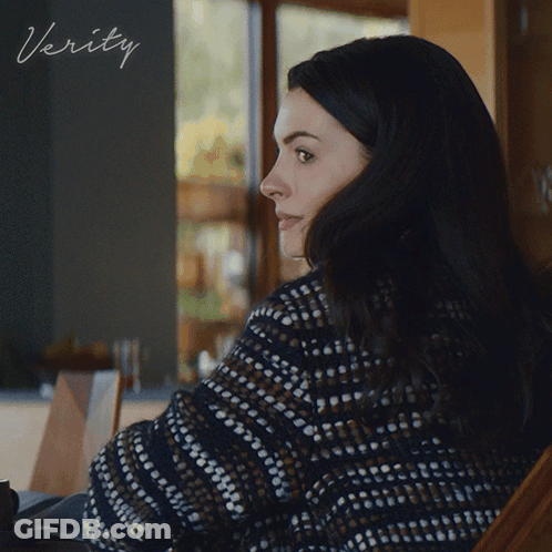 A Woman In A Sweater Sits In Front Of A Sign That Says ' Verity ' On It GIF