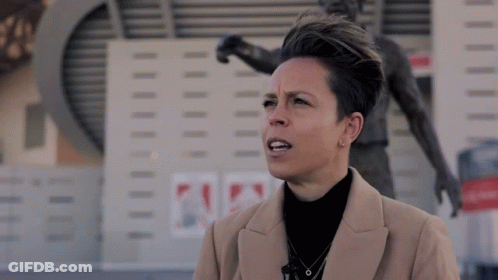 A Woman In A Tan Coat Stands In Front Of A Statue Of A Soccer Player GIF