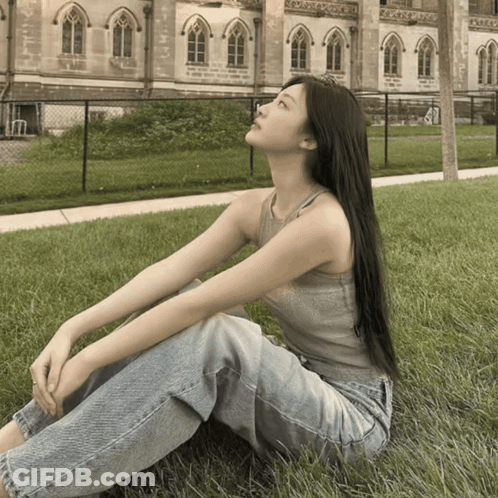 A Woman In A Tank Top And Jeans Is Sitting In The Grass GIF