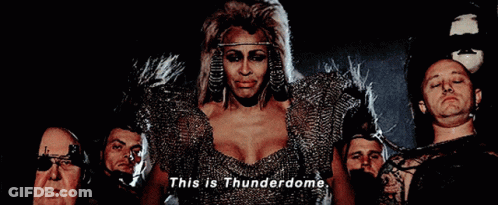 A Woman In A Thunderdome Costume Is Surrounded By Men GIF