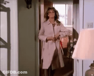 A Woman In A Trench Coat Is Standing In A Hallway . GIF
