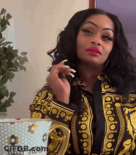 A Woman In A Versace Shirt Is Sitting At A Table With A Cup . GIF
