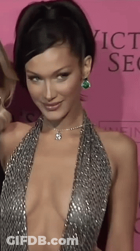 A Woman In A Very Plunging Dress Is Standing In Front Of A Pink Background That Says Victoria 'S Secret GIF