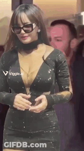 A Woman In A Very Revealing Black Dress And Sunglasses Is Standing In Front Of A Man . GIF