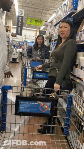A Woman In A Walmart Shopping Cart With A Sign That Says Attention Shoppers On It GIF