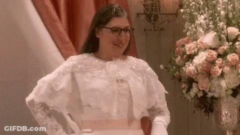 A Woman In A Wedding Dress And Gloves Is Standing Next To A Vase Of Flowers . GIF