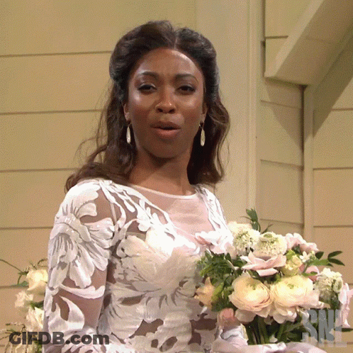 A Woman In A Wedding Dress Holds A Bouquet Of Flowers In Front Of A Sign That Says Nbc GIF