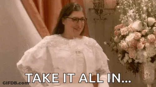 A Woman In A Wedding Dress Is Standing In Front Of A Vase Of Flowers And Says `` Take It All In '' . GIF