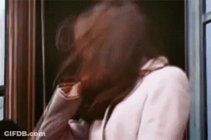 A Woman In A White Coat Is Standing In Front Of A Window Holding Her Hair . GIF