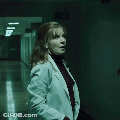 A Woman In A White Coat Stands In A Hallway GIF