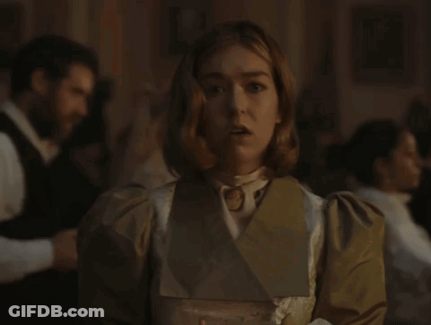 A Woman In A White Dress And A Brown Jacket Stands In A Dark Room GIF