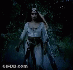 A Woman In A White Dress And A Horned Hat Is Walking Through A Dark Forest . GIF