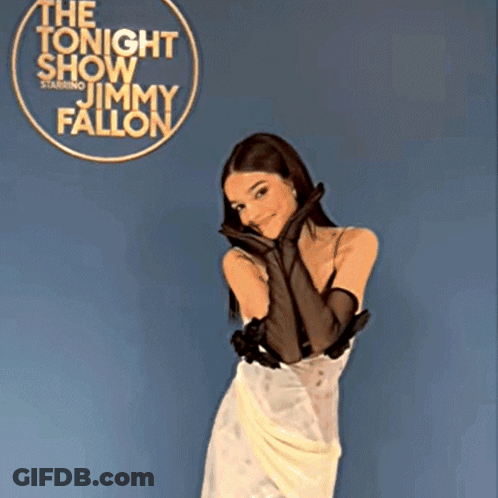 A Woman In A White Dress And Black Gloves Stands In Front Of A Sign For Jimmy Fallon GIF