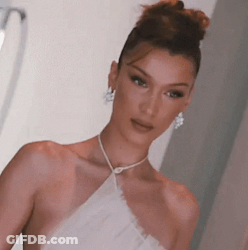 A Woman In A White Dress And Earrings Is Standing In A Room . GIF