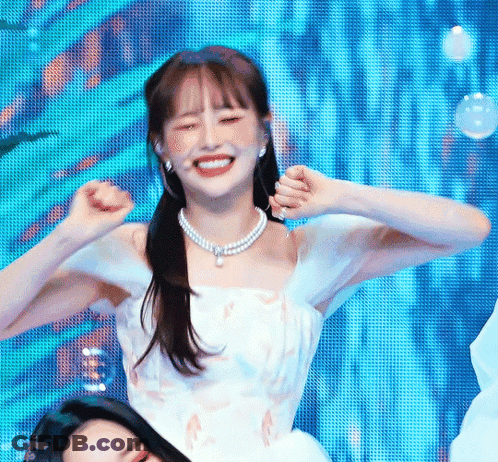 A Woman In A White Dress And Pearl Necklace Is Dancing On A Stage With Her Hands In The Air . GIF