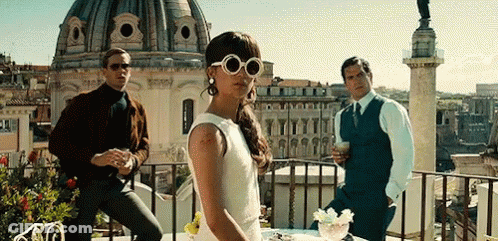 A Woman In A White Dress And Sunglasses Is Standing On A Balcony Next To Two Men . GIF