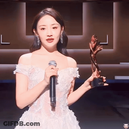 A Woman In A White Dress Holding A Microphone And A Trophy GIF