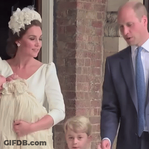 A Woman In A White Dress Holds A Baby While A Man In A Suit Looks On GIF