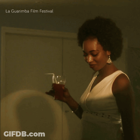 A Woman In A White Dress Holds A Glass In Front Of A La Guarimba Film Festival Poster GIF