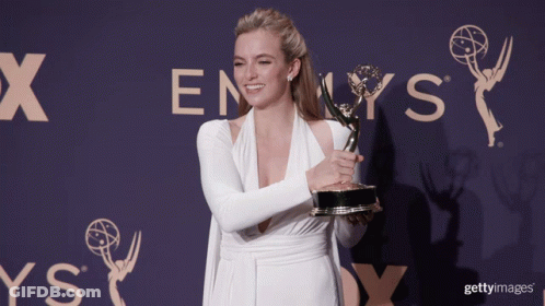 A Woman In A White Dress Holds A Trophy In Front Of An Emmys Sign GIF