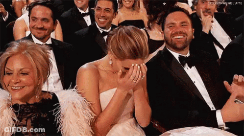 A Woman In A White Dress Is Covering Her Face With Her Hands While Sitting In A Crowd Of People . GIF