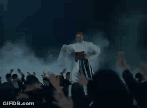 A Woman In A White Dress Is Dancing In Front Of A Crowd Of People While Flames Are Coming Out Of Her Dress . GIF
