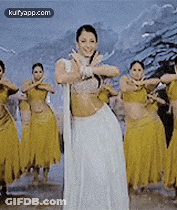 A Woman In A White Dress Is Dancing In Front Of A Group Of Dancers In Yellow Skirts . GIF