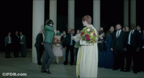 A Woman In A White Dress Is Holding A Bouquet Of Flowers In Front Of A Crowd Of People GIF