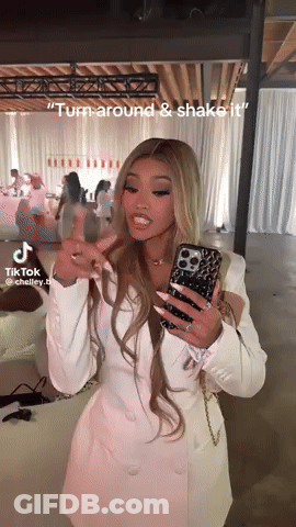 A Woman In A White Dress Is Holding A Cell Phone In Her Hand And Says " Turn Around & Shake It " GIF