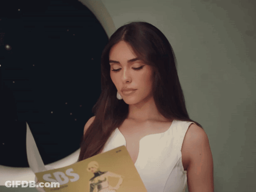 A Woman In A White Dress Is Holding A Magazine That Says Sbs GIF
