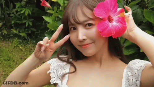 A Woman In A White Dress Is Holding A Pink Flower In Front Of Her Face GIF