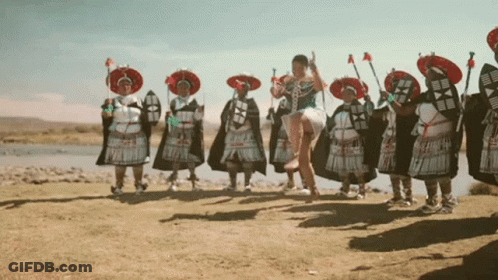A Woman In A White Dress Is Jumping In The Air In Front Of A Group Of People In Traditional Costumes GIF