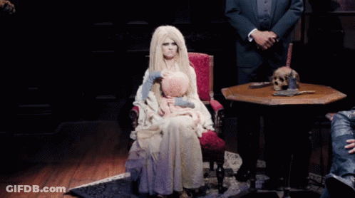 A Woman In A White Dress Is Sitting In A Chair With A Doll On Her Lap GIF