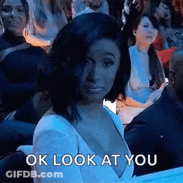 A Woman In A White Dress Is Sitting In A Crowd Of People And Looking At The Camera . GIF