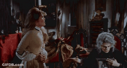 A Woman In A White Dress Is Standing In Front Of A Bed With Red Sheets GIF