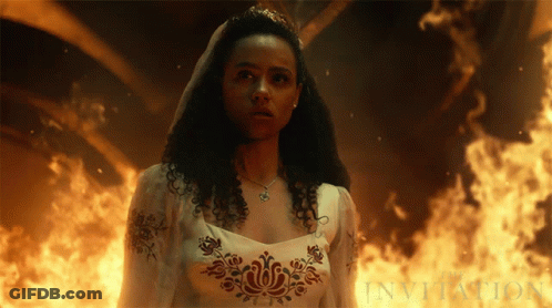 A Woman In A White Dress Is Standing In Front Of A Fire And The Word Invitation Is Visible GIF