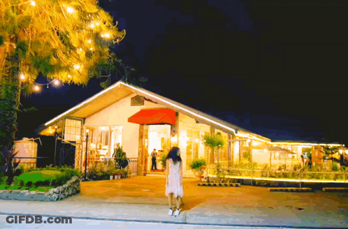 A Woman In A White Dress Is Standing In Front Of A House With A Red Awning GIF
