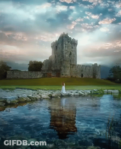 A Woman In A White Dress Is Standing In Front Of A Large Stone Castle GIF